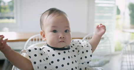 A baby is sitting in a high chair with a polka dot shirt on. The baby is looking at the camera with...