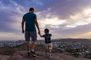 infant holding father hand and exploring nature at mountain top with dramatic sky and city view