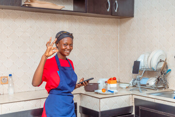 Excited Young Black Female Cook Using Mobile Phone and Gesturing Okay in Kitchen