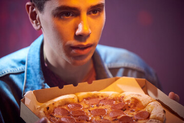Young calm guy with pepperoni pizza in a cardboard box in the evening in a pizzeria