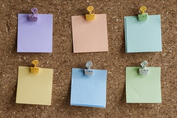 Six blank pastel-colored post-it notes are pinned neatly to a brown cork board
