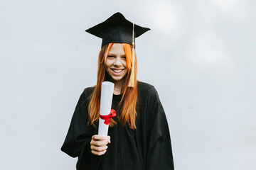 teenage girl in clothes of a graduate coat and cap celebrates high school or  junior year graduation on background of white wall with shadows and with diploma in hands, education and no school concept