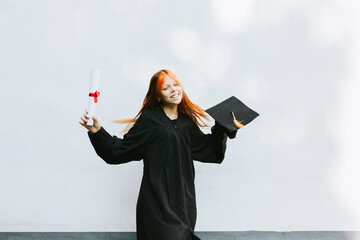 teenage girl in clothes of a graduate coat and cap celebrates high school or  junior year graduation on background of white wall with shadows and with diploma in hands, education and no school concept