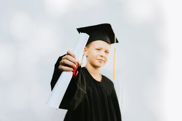 teenage boy in clothes of a graduate coat and cap celebrates high school or  junior year graduation on background of white wall with shadows and with diploma in hands, education and no school concept