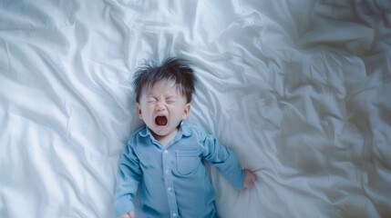 A young child in a blue shirt lying on a white bed, crying with eyes closed and mouth open.
