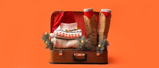 Suitcase with Santa hat, warm clothes, Christmas balls and fir branches on orange background....