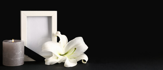 Beautiful lily flower with blank funeral frame and burning candle on dark background