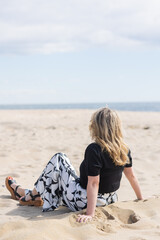 Back of blonde woman in black top, black and white graphic print pants and black sandals sitting in...