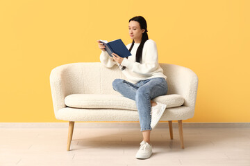Beautiful young woman with book sitting on comfortable sofa near yellow wall
