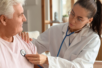 Young nurse hearing senior man with stethoscope at home