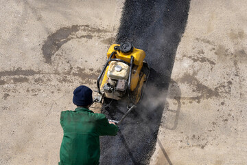 A road service worker compacts asphalt with a mechanical compactor.	