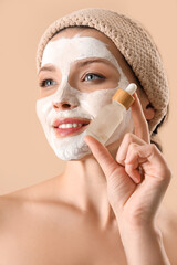 Beautiful woman with applied facial mask and bottle of serum on beige background, closeup