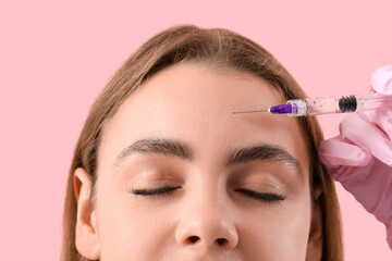 Young woman receiving filler injection in forehead on pink background