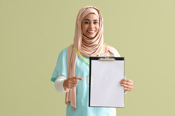 Female Muslim medical intern pointing at clipboard on green background
