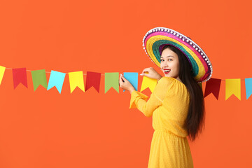 Happy young woman in Mexican sombrero hat and with festive garland on orange background