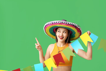 Happy young woman in Mexican sombrero hat and with garland pointing at something on green background