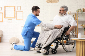Young caregiver covering senior man in wheelchair with warm plaid at home