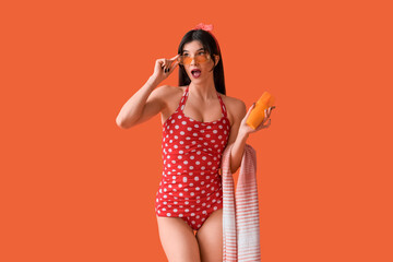 Beautiful young shocked woman in swimsuit with bottle of sunscreen cream on orange background....