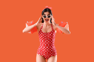 Beautiful young shocked woman in swimsuit with inflatable armbands on orange background. Summer vacation concept
