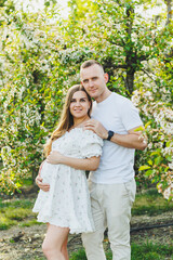 Fototapeta premium Tender romantic pregnant couple hugging in blooming apple spring garden. Expectant parents in the park of white flowering trees. A couple is expecting a baby