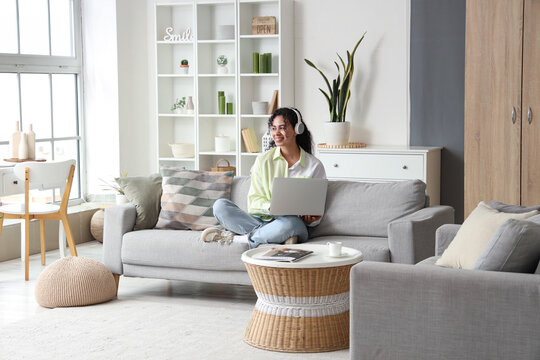 Young African-American woman in headphones using laptop on sofa at home