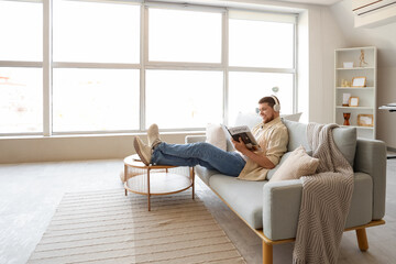 Young bearded man in headphones reading magazine on sofa at home