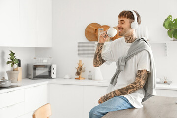 Young tattooed man with headphones and coffee cup listening to music in kitchen