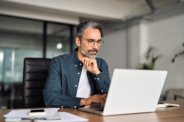 Busy middle aged business man executive wearing shirt and glasses sitting at desk using laptop....