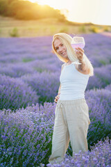 Lavender field. Beautiful woman with blond hair walking between rows of lavender flowers with glass of summer refreshing cocktail, relaxing, resting, enjoying nature. Mental health concept