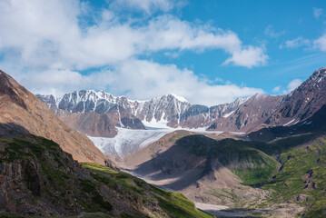 Scenic landscape with glacier tongue among rocky ridges and sharp snow-capped mountain range in...