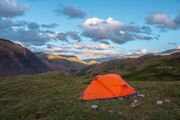 Vivid orange tent on grassy hill with view to mountain tops illuminated by sunset light. Rocks...