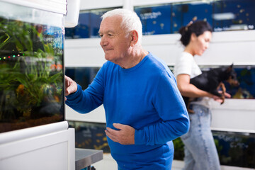 Old man standing in pet shop and looking at aquarium and choosing fishes. Woman with dog in hands standing in background.