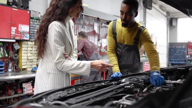 Young auto mechanic explaining to his female customer what needs to be fixed in her car, while his colleague working in the background