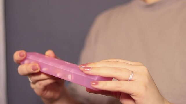 Close up of a woman taking a pill out of a pink pillbox, blurred background 