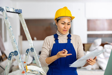 Disappointed Asian woman worker checking documents during repair works indoors.