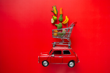  car with a shopping basket on the roof with fruits and vegetables on a red background.Delivery of...