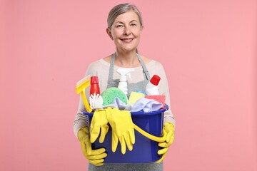 Happy housewife holding bucket with cleaning supplies on pink background