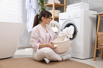 Happy young housewife putting laundry into washing machine at home