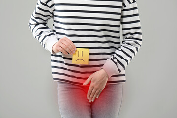 Woman holding sticky note with drawn sad face and suffering from cystitis symptoms on light grey background, closeup