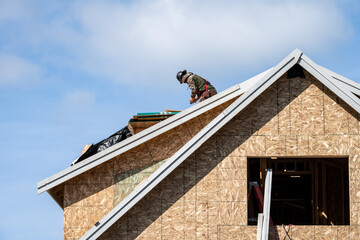 Workman in a hard hat and safety harness working on roof of a new home under construction
