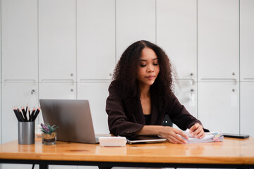 Focused African American woman sorting through paperwork at her office desk, with a laptop and smartphone nearby.