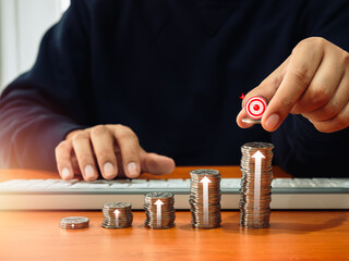 Businessman hand put 3d target icon on coins stacks as graph steps with rising arrows on desk while...