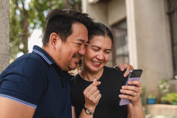 Middle-aged Southeast Asian husband and wife watching a video greeting from a friend. Making a finger heart gesture.