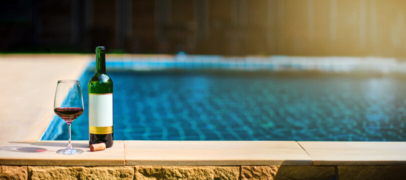 A bottle of red wine and a glass of wine are set on the edge of a clear blue pool, surrounded by a lush green lawn. The afternoon sun shines down, creating a relaxing and inviting atmosphere.