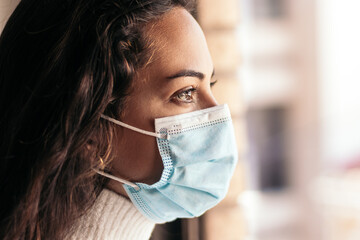 Young woman wearing face mask looking out the window at home