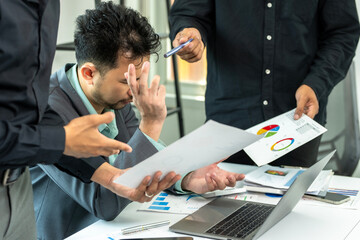 A man is sitting at a desk with a laptop and several papers in front of him. He is frustrated and is holding a pen
