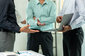 Three men are standing around a table with a laptop and papers. They are discussing something important. Scene is serious and focused