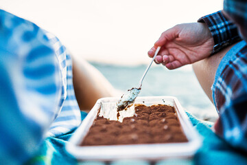 A close-up view of a hand scooping a portion of tiramisu from a container, with the backdrop of a...