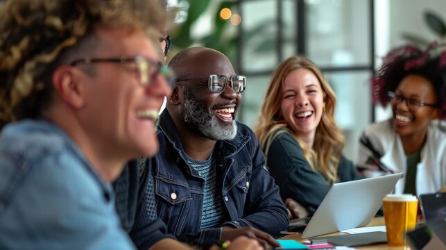 A group of diverse colleagues sitting around a table in a modern office. They are all smiling and laughing, and they look like they are enjoying each other's company. 