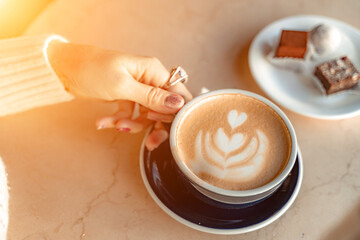 A cup of fresh cappuccino coffee in the hands of a woman on a fashionable background of a white marble table, next to a plate with sweets. Coffee addiction. Top view, flat lay.